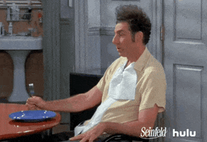 Seinfeld gif. Michael Richards as Kramer gazes forward at a table and idly taps his foot and his fist in time, holding a fork.