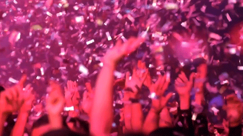 Pohyblivý obrázek s davem lidí vztahujícími ruce vzhůru a chytajícími padající konfety na koncertě. 