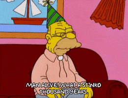 The Simpsons gif. Grandpa sits on a couch with a birthday hat on his head. He angrily says, “Man alive! What a stinko thousand years.” 