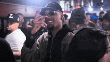 Video gif. Man stands in a crowd and wears a swaggy fit with black shades, a black cap, and an army green jacket on top of a black zip up jacket. He holds up his hand in front of his face and spreads his fingers to casually gesture, "What's up?"