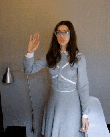 Video gif. We follow a hand as it high-fives a woman in a gray dress with pointy blue-rimmed glasses.