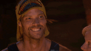 Reality TV gif. David Genat grins wide on Australian Survivor while smoke drifts in the background.