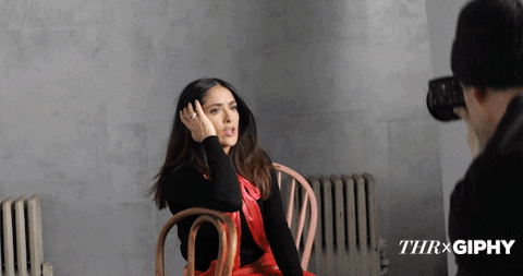 A light-skinned woman posing for a photograph whilst sitting on a brown chair. She's holding her hair and pushing it back with one arm
