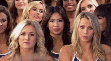 Reality TV gif. A contestant on Dallas Cowboys Cheerleaders: Making the Team gasps and gestures toward herself in surprise as the women around her clap and turn to look at her.