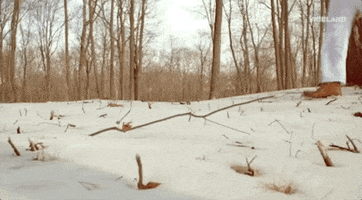 Video gif. From a ground view, we see the legs of a person wearing white pants and brown boots walking through a snowy forest, kicking up snow as they walk.