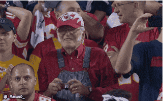 Video gif. Standing in the audience of a football game, an old man wearing overalls and a Kansas City Chiefs hat bites his bottom lip as he squints out at the field. He then holds up a small digital camera up to his face and squints into it to take a picture.