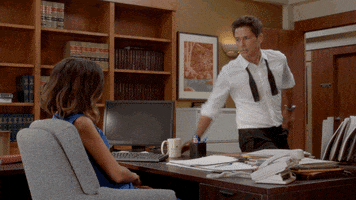 TV gif. Rob Lowe as Dean Sanderson on The Grinder wipes everything off of a woman’s messy office desk. The woman sits in her chair unamused, but Dean stands across from her with his tie undone. 