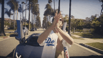 Celebrity gif. Wearing an unbuttoned Dodgers jersey, Rapper Lil Dicky stands in front of a Lamborghini, clapping his hands enthusiastically.
