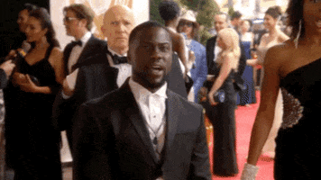 TV gif. Kevin Hart on Real Husbands of Hollywood stands on a red carpet among well-dressed celebrities, and recoils with shock when someone tosses something at him that stains his suit jacket yellow.