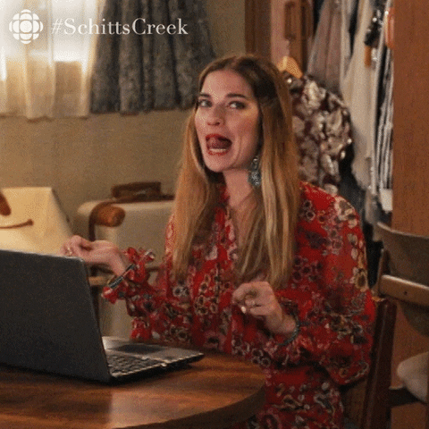 Schitt's Creek gif. A giddy Annie Murphy as Alexis is seated in front of a laptop at a small table. While looking at us, she touches her tongue to her upper lip and playfully tosses her hair around.