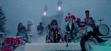 Music video gif. Elton John plays the piano and Ed Sheeran plays the guitar in the Merry Christmas music video. Snow falls around them and their band members. There’s a castle in the background.
