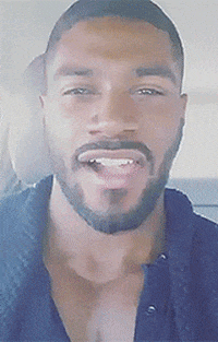 Video gif. Man sits in a car and looks at us with a flirtatious smile as he slowly chews on gum.