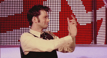 Celebrity gif. David Tennant sits and looks to his right, clapping.