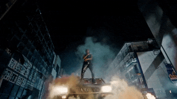 Celebrity gif. In the Car Bandana music video, Fireboy DML stands atop a black car in the nighttime city streets and sings as flames and smoke billow around him.