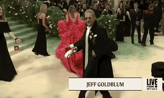 Met Gala 2024 gif. Jeff Goldblum strides across the red carpet wearing a long black overcoat, black straight leg trousers, and high-shine square toed shoes. He's holding white gloves and wearing a collection of diamond floral-shaped brooches on his lapel. 