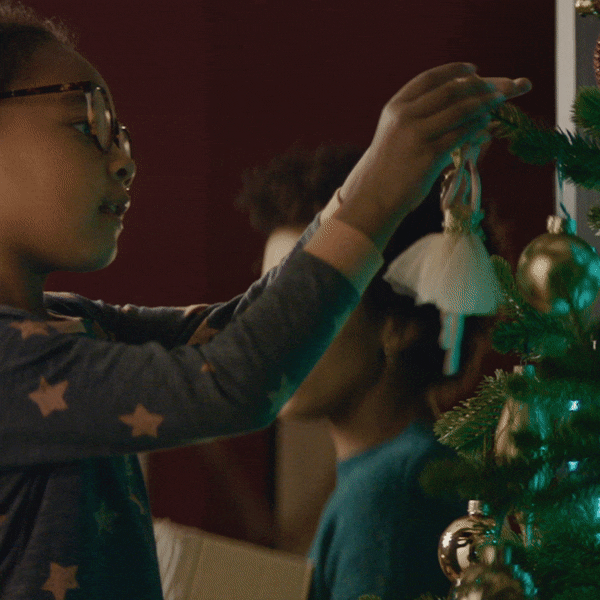 Holiday gif. Young girl in star-decorated pajamas delicately places a ballerina ornament on the Christmas tree.