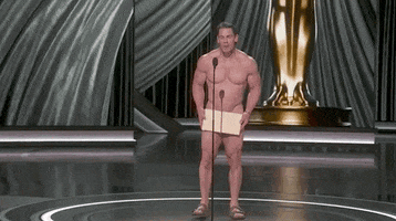 Oscars 2024 GIF. A naked John Cena has finally made it to the microphone with the Oscars envelope covering his privates. He shakes his head with the tremendous effort its taken to get there and he says, "Costumes." 