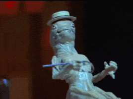 Movie gif. Small alien from Spaceballs wears a hat and holds a cane. He kicks his legs up high as he bobs its small alien head side to side.