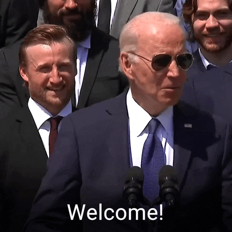 Political gif. Joe Biden wearing sunglasses and standing behind microphones saying, "welcome."