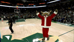 Sports gif. Boston Celtics mascot struts across the basketball court in a Santa costume, waving his arms to rev up a stadium full of fans. 