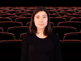 Digital compilation gif. Young woman claps her hands in an exaggerated way then leans forward at us as popcorn pops up into the air all around her in front of a movie theater background. Text, "The movie was better."