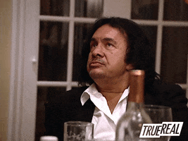 Reality TV gif. Gene Simmons on Family Jewels sits at a dining table. He looks up, rolling his eyes, in irritation.