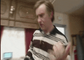 Video gif. Taking himself way too seriously, a dorky man plays air guitar in his living room.
