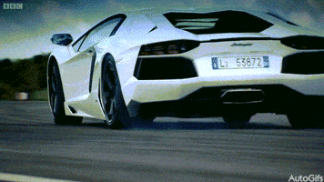 Video gif. From behind, a white Lamborghini Aventador drifts gently on the road before catching traction and turning.