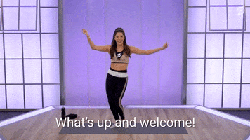 Video gif. A Peloton instructor standing on a yoga mat raises up her arms in excitement as she says, “What’s up and welcome!”