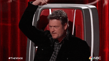 TV gif. Blake Shelton as a judge on the Voice sits in a high-backed red chair pointing his index finger down at the top of his head.