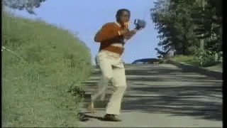 Video gif. A man in an orange and striped sweater and beige flared pants dances down a tree-lined street singing on a blue sunny day, microphone in one hand and radio in the other. 