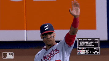 Sports gif. Juan Soto from the Washington Nationals is on the field and gives us a high wave with a serious expression as he chews a piece of gum. 