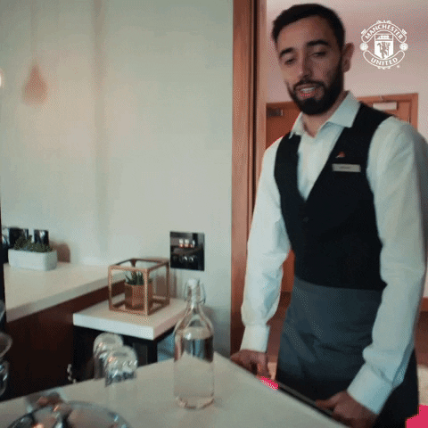 Video gif. Bruno Fernandes from Manchester United wearing a vest and service worker's name tag rolls a cart with glasses into a kitchen as he looks at us with a serene expression and says, "How may I assist?'