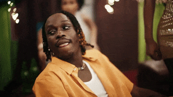 Music video gif. Fireboy DML in the Bandana music video sits in a chair with his legs spread out. Women stand around him in dresses. He looks around at the women and smirks at us.