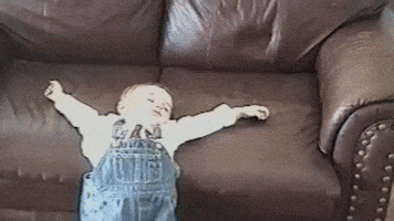 Video gif. A toddler is passed out half on and half off a couch. Their arms are splayed fully outwards and they look sloshed.