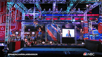 Reality TV gif. A man on American Ninja Warrior slips off a monkey bar on a wheel and falls into a pool of water as the audience and a family member on a screen look on. 