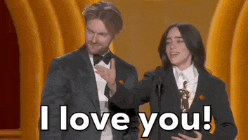 Oscars 2024 gif. Billie Eilish and Finneas O'Connell win Best Song. With her hand, Eilish gestures towards someone in the crowd and says passionately, "I love you!" O'Connell smiles in agreement. 