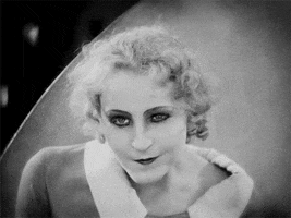 Black and white gif of Maria, seen as a white woman with curly hair. She smiles slyly and raises one eyebrow.