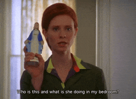 TV gif. Cynthia Nixon as Miranda Hobbes on Sex and The City has a very serious and irritated expression on her face as she holds up a small statue of Mother Mary. She says, “Who is this and what is she doing in my bedroom?”