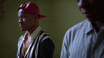 Movie gif. Tequan Richmond as Gabe in Savage Youth looks at something off screen with a slight smile before he bursts out laughing and leans forward with his arms crossed over his stomach. 