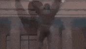 Movie gif. Sylvester Stallone as Rocky Balboa in the movie, "Rocky" raises his fists over his head as he bounces up and down in victory against a smoggy city background.