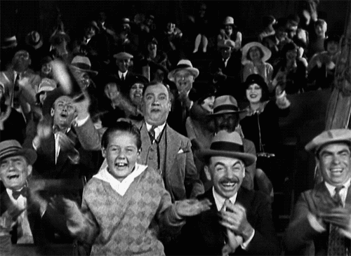 Movie gif. In the style of an old movie, a large crowd of people dressed in 1930s-era clothes cheer and whoop and holler wildly. Maybe someone just invented sliced bread?