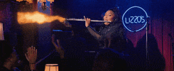 Celebrity gif. Musician Lizzo in a black sequin bodysuit enthusiastically plays a flaming flute to an applauding audience. 