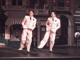 Movie gif. Gene Kelly and Frank Sinatra in Take me Out to The Ball Game wear matching striped suits. In unison, they dance, clicking their heels in the air with their hands in their pockets.