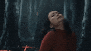 Music video gif. Mitski is in a fiery forest and looks as if she's in pain. She throws a hand out and moves towards us jerkily, as if she's being summoned and cannot resist.