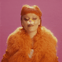 Video gif. Woman wearing an orange lion nose, furry orange jacket, and an orange wig with bangs leans in and smiles at us, giving two thumbs up against a dark pink background. Text, "Double thumbs up."