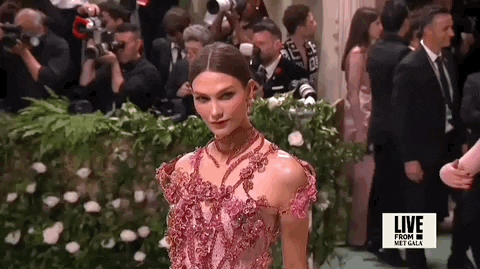 Met Gala 2024 gif. Close up of the top of Karlie Kloss wearing a jeweled pink floral Swarovski gown with ropes of fabric weaving an intricate design across her torso and around her neck. 