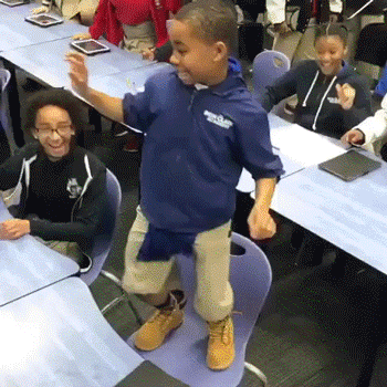 Video gif. In a classroom full of dancing children, a happy child in a blue hoodie dances while standing on a chair. Impressed, the child next to him stands up and dances along.