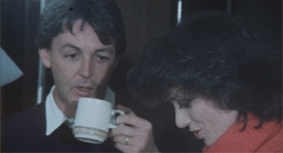 Celebrity gif. Paul McCartney sips from a white cup and gives a side-eyed stare at a woman in a red turtleneck.