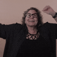 Reaction gif. A Disabled white woman with kinky curly gray hair and big wine-colored cat-eye glasses waves her arms above her head in a rhythmic happy dance, ending with a holler and a cheer.